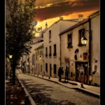 Rue pavée au coucher du soleil, bordée de bâtiments anciens avec des lampadaires allumés, ornée de street art et traversée par deux passants.