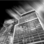 Photographie en noir et blanc de gratte-ciels modernes vus en contre-plongée, avec un effet de mouvement dans le ciel.