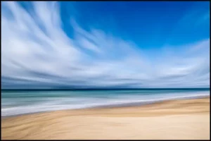Paysage côtier avec une plage dorée et un océan aux teintes turquoise, sous un ciel bleu dynamique traversé par des nuages en mouvement.