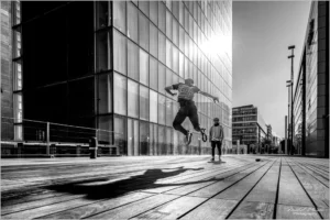 Un skateur en plein saut réalise une figure aérienne sur une esplanade urbaine, projetant une ombre dynamique sur le sol en bois, tandis qu’un ami observe la scène devant des immeubles modernes vitrés
