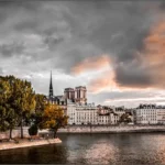 Vue sur les rives de la Seine à Paris avec Notre-Dame en arrière-plan, sous un ciel dramatique où les nuages sombres contrastent avec des teintes dorées de coucher de soleil