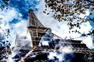 Vue artistique et dynamique de la Tour Eiffel, capturée en contre-plongée avec un effet de réflexion et de superposition de nuages et de feuillage.