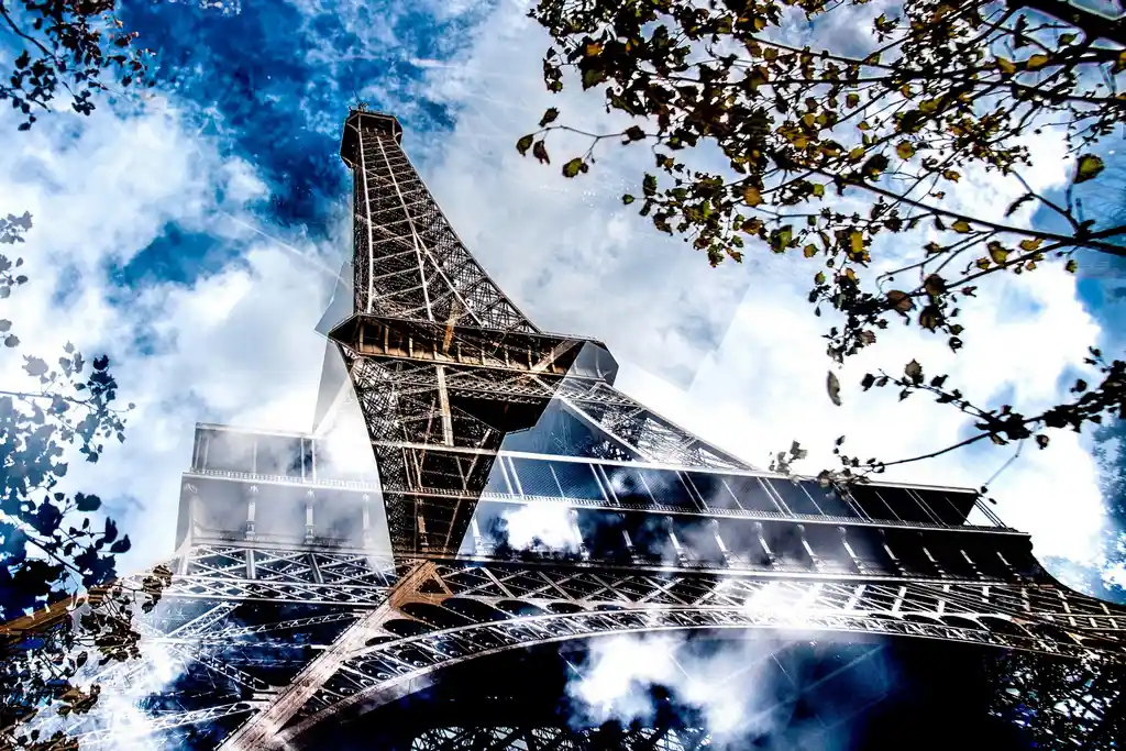 Vue artistique et dynamique de la Tour Eiffel, capturée en contre-plongée avec un effet de réflexion et de superposition de nuages et de feuillage.