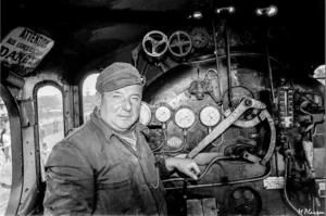 Portrait en noir et blanc d’un conducteur de locomotive à vapeur dans sa cabine, entouré de cadrans, leviers et manomètres